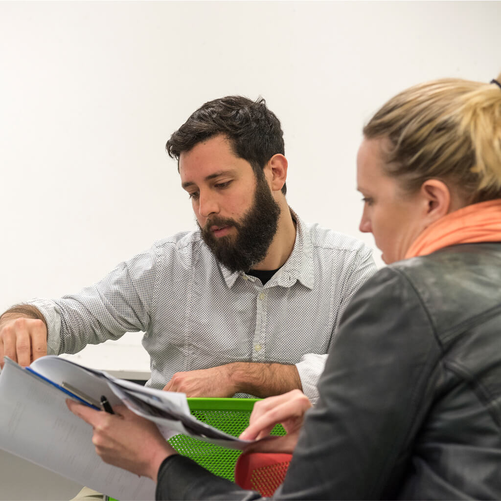 Un homme et une femme examinent ensemble des documents à un bureau ; l'homme fait un geste vers une page tandis que la femme, qui discute peut-être d'art-thérapie, tient les papiers.