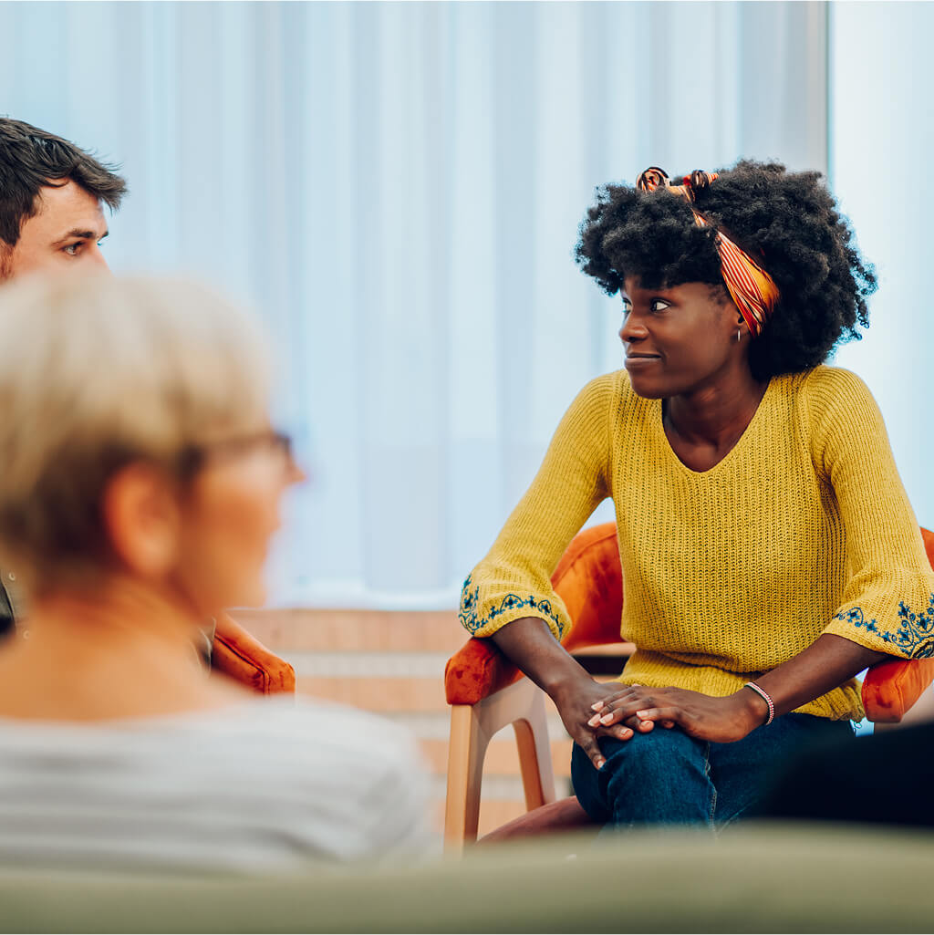 Une femme vêtue d'un pull jaune est assise et écoute une discussion de groupe sur la santé et le bien-être avec trois autres personnes dans une salle bien éclairée.