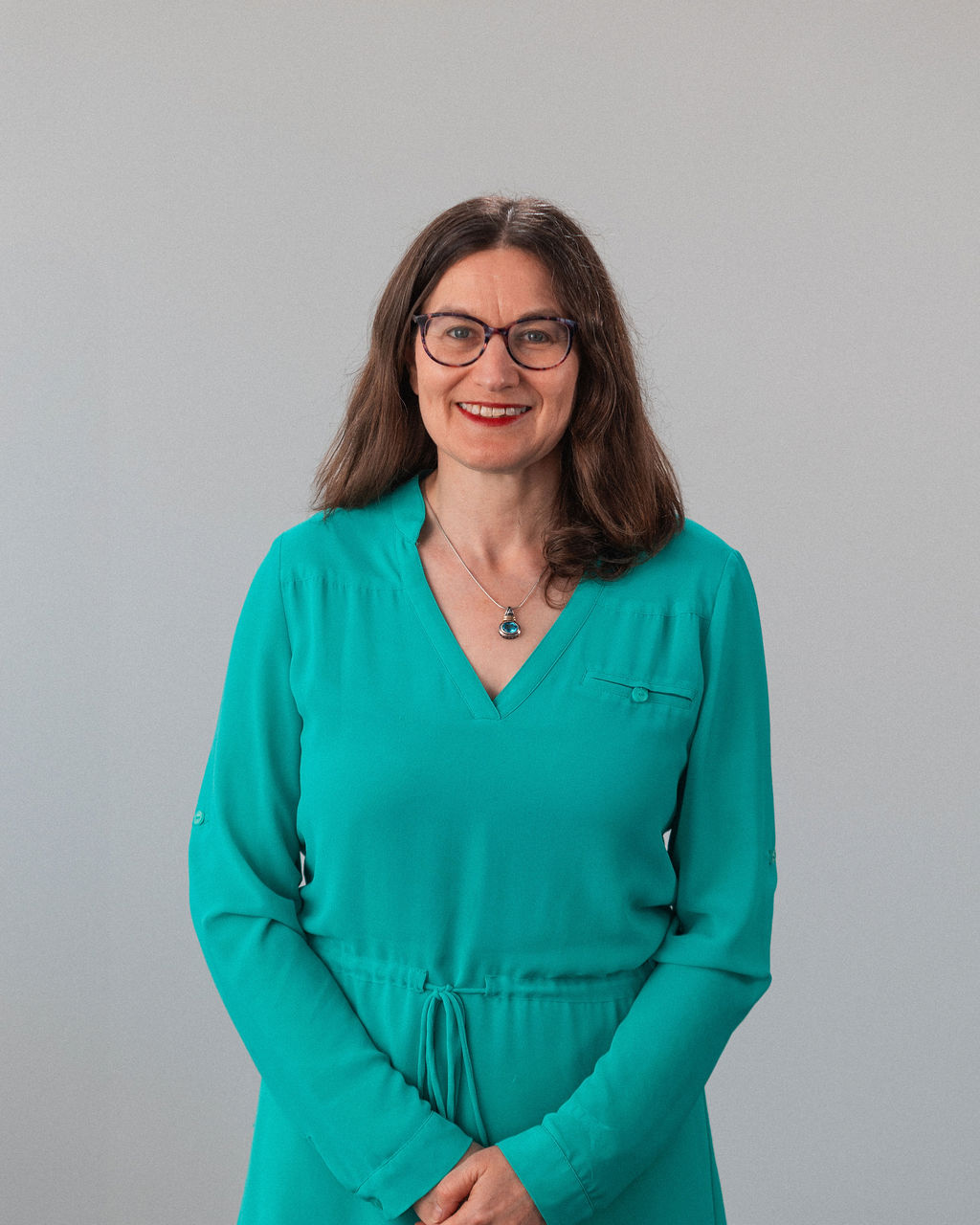 Nicole Dorfan, une femme aux longs cheveux bruns, portant des lunettes et une robe sarcelle, se tient sur un fond clair, souriante, les mains croisées devant elle.