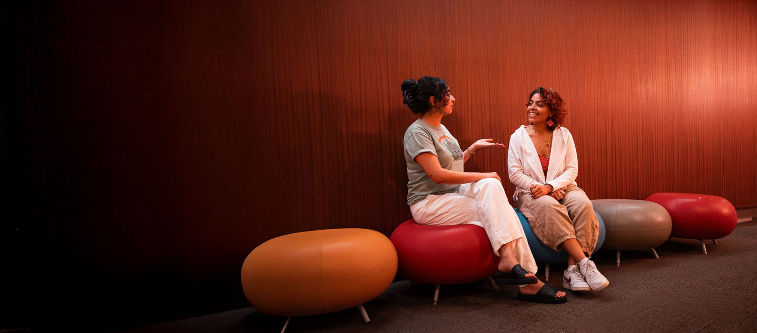 Dos estudiantes de la Universidad Adler conversan sentados en taburetes redondos de colores.