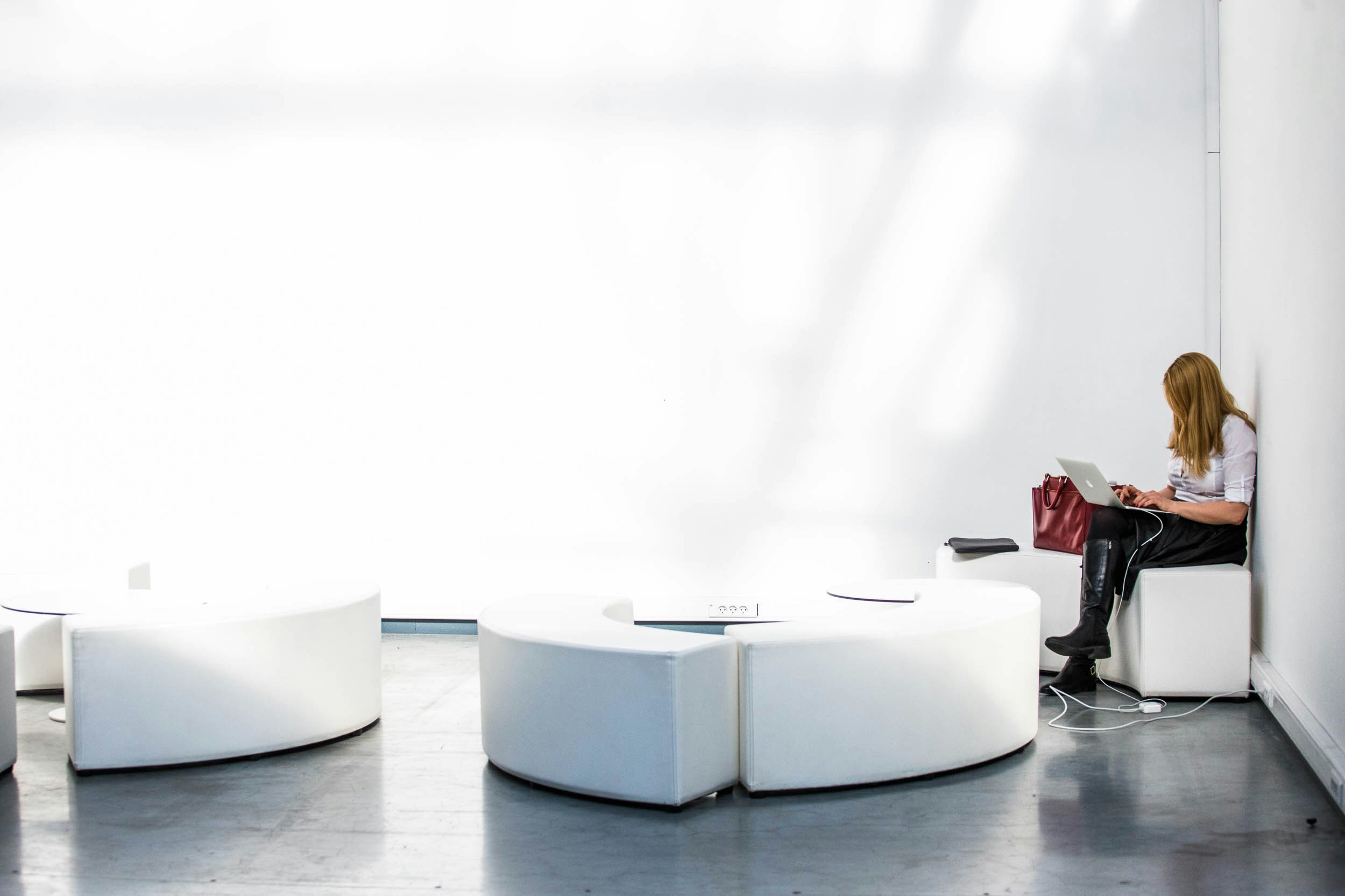 A woman sits alone on a white curved bench, working on a laptop with a red bag next to her in a bright, minimalist room.
