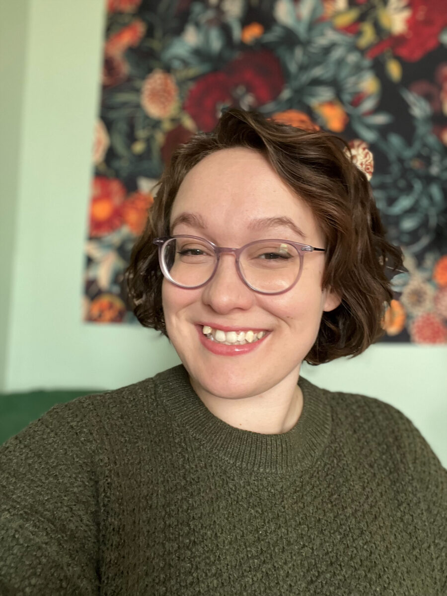 Una persona sonriente con el pelo rizado y gafas lleva un jersey verde. Un colorido tapiz floral cuelga de fondo, creando un ambiente alegre.