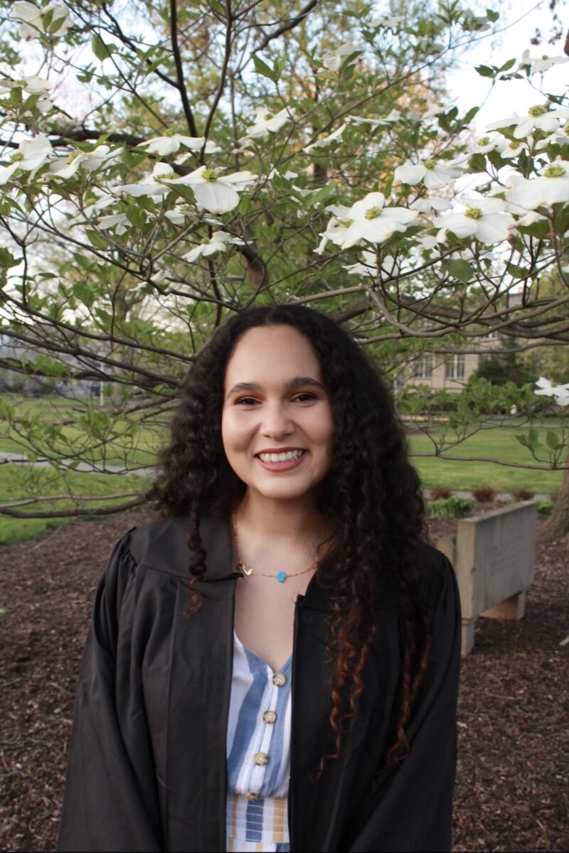Una joven de pelo rizado sonríe cálidamente, vestida con un traje de graduación. Está delante de un árbol en flor, que transmite felicidad y éxito.