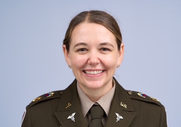Una persona con uniforme militar estadounidense e insignias médicas sonríe a la cámara sobre un fondo azul claro liso, identificada como Amy Thrasher, Psy.D.