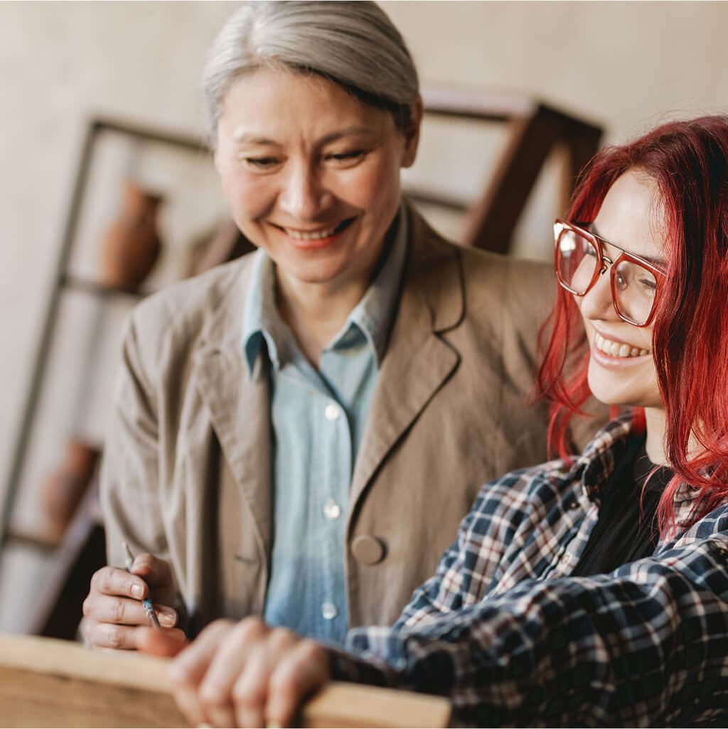Deux femmes se tiennent ensemble à l'intérieur, l'une avec des cheveux gris et une veste beige, l'autre avec des cheveux roux et des lunettes, toutes deux souriantes alors qu'elles discutent d'études de maîtrise en art-thérapie ou en psychologie de l'orientation.