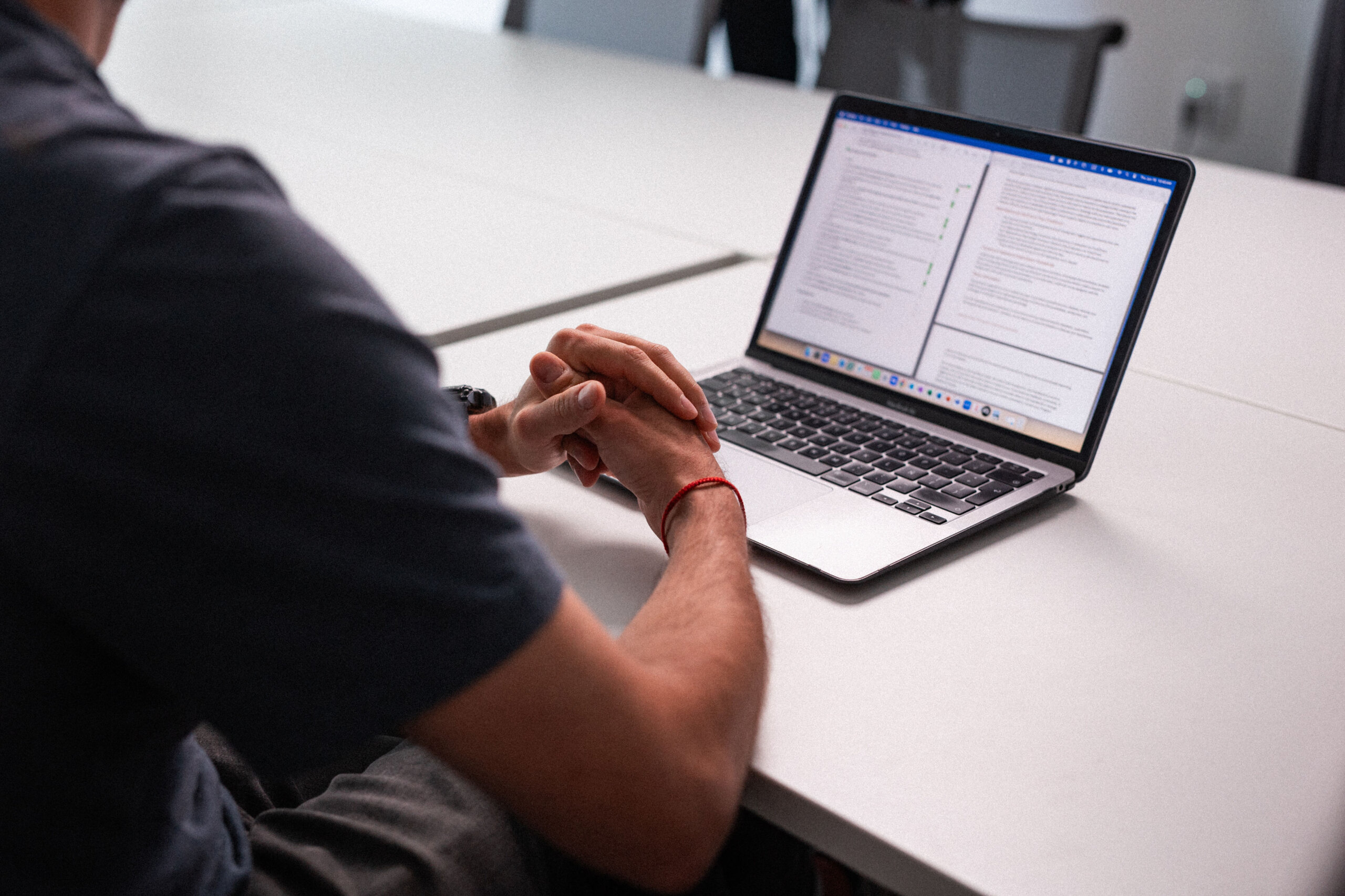Person sitting at a white table with hands clasped, looking at an open laptop displaying documents with text and reviewing Website Requests.