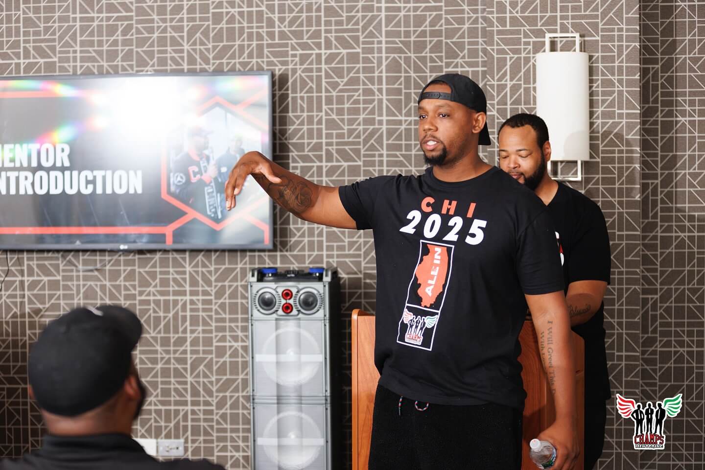Un hombre con una camiseta de "CHI 2025", antiguo alumno de Adler, habla de cómo la tutoría transforma vidas a un público sentado, mientras otro hombre se sitúa detrás de él en un podio. En una pantalla se lee "Mentor Introduction.