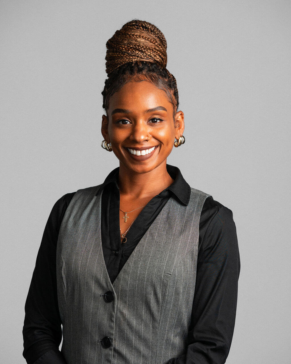 Dominique Barnes-Walker, une femme aux cheveux tressés en chignon haut, vêtue d'une chemise noire et d'un gilet gris, sourit à l'appareil photo sur un fond gris uni.