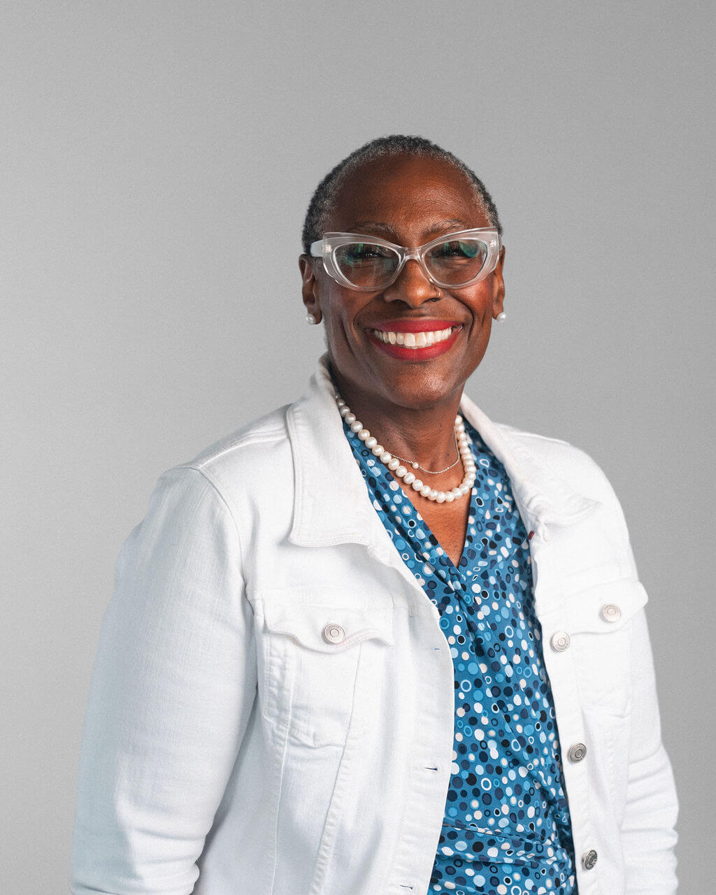 Monica Boyd-Layne, une femme âgée portant des lunettes, une veste blanche, un chemisier bleu à motifs et un collier de perles, sourit chaleureusement sur un fond gris uni.