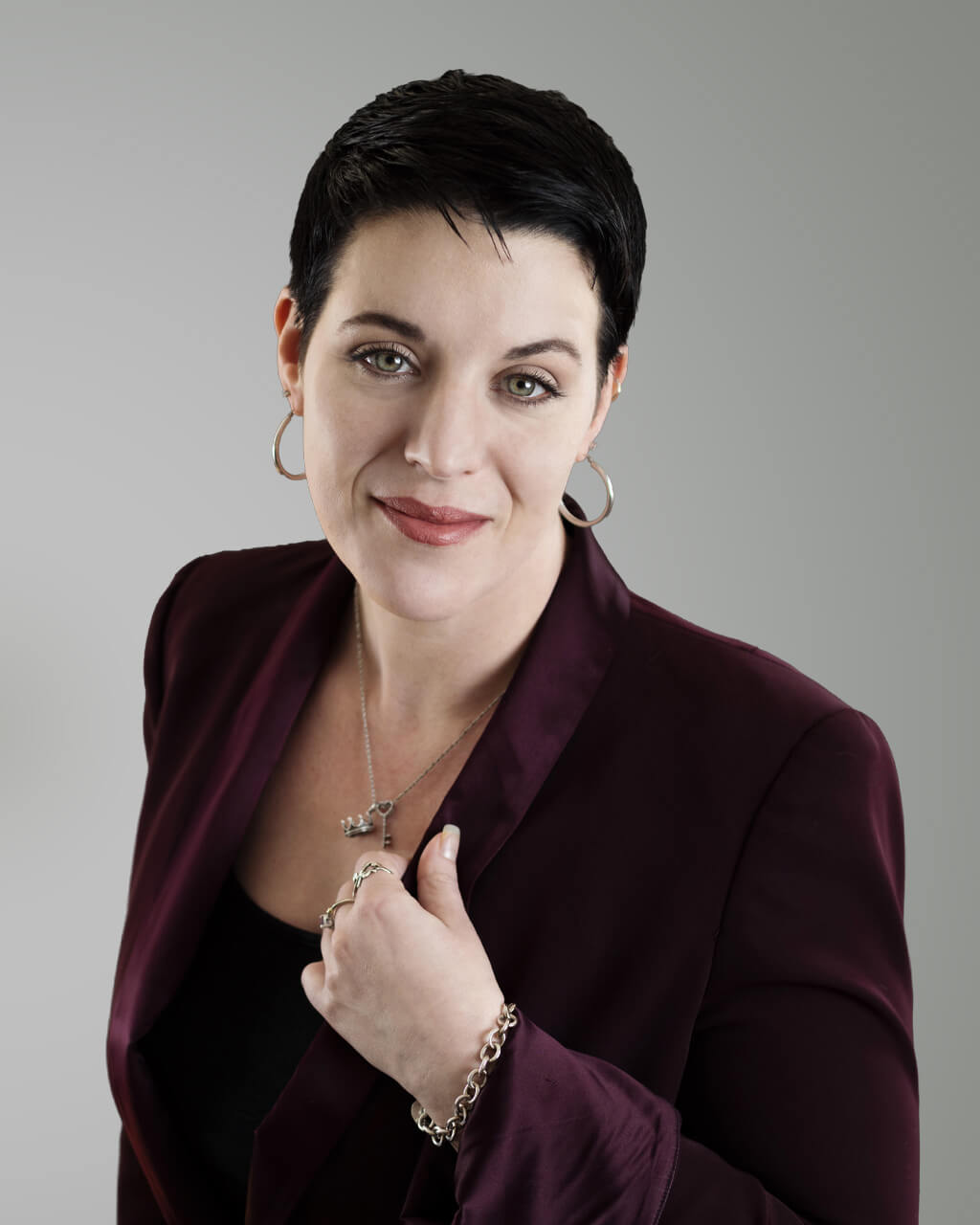 Karen Caffee, une femme aux cheveux courts et foncés, se tient avec assurance devant un fond gris uni. Elle porte un blazer bordeaux par-dessus un haut noir, rehaussé de bijoux en argent et de boucles d'oreilles créoles.